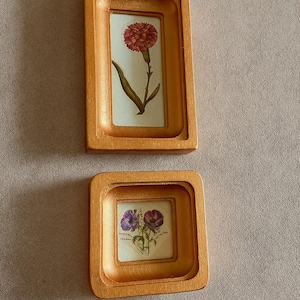 May include: Two small, gold-colored framed botanical prints. The top frame is rectangular and contains a red flower. The bottom frame is square and features purple flowers. The frames are set against a neutral background.
