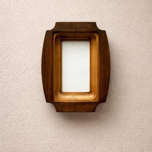 Op de afbeelding: Een rechthoekige houten fotolijst met een lichtbruine afwerking. De lijst heeft een verzonken binnenrand en een wit interieur. De lijst staat tegen een getextureerde, lichtroze achtergrond.