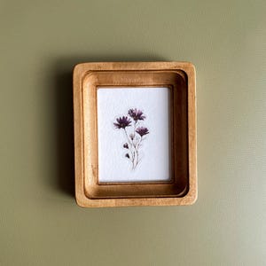 以下が含まれることがあります： 金色の仕上げが施された、小さな長方形の木製フレーム。フレーム内には、紫色の花のクラスターが描かれた白い紙が入っています。フレームは、くすんだ緑色の背景に配置されています。
