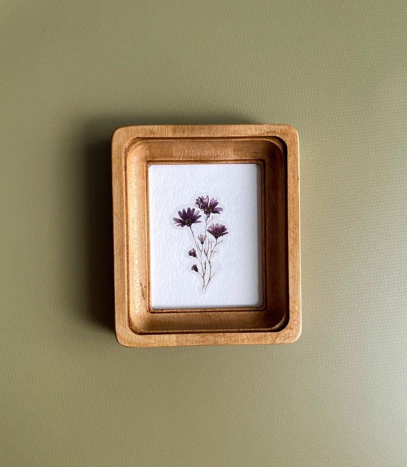 以下が含まれることがあります： 金色の仕上げが施された、小さな長方形の木製フレーム。フレーム内には、紫色の花のクラスターが描かれた白い紙が入っています。フレームは、くすんだ緑色の背景に配置されています。