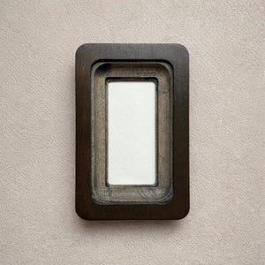 May include: A rectangular dark brown wooden frame with rounded corners and a recessed inner border. The frame features a smooth, polished finish with visible wood grain patterns and a centered, plain white rectangular insert for display.
