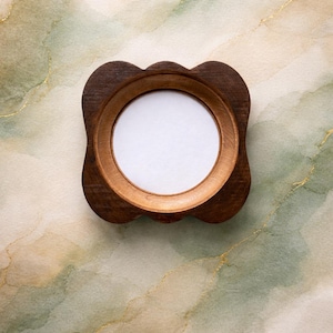 May include: A small, round wooden picture frame with a dark brown finish. The frame has a scalloped edge and a light brown interior. The center of the frame is white, ready for a photo or artwork. The background is a watercolor wash of green, gold, and cream.