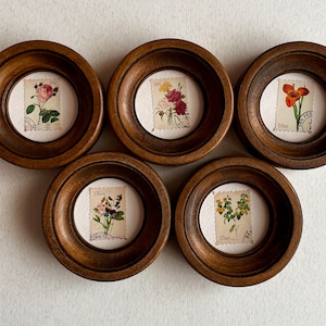 Peut inclure: Cinq cadres ronds en bois avec des faces en verre, chacun affichant un timbre floral vintage. Les timbres présentent différentes fleurs et ont des prix variés imprimés dessus.