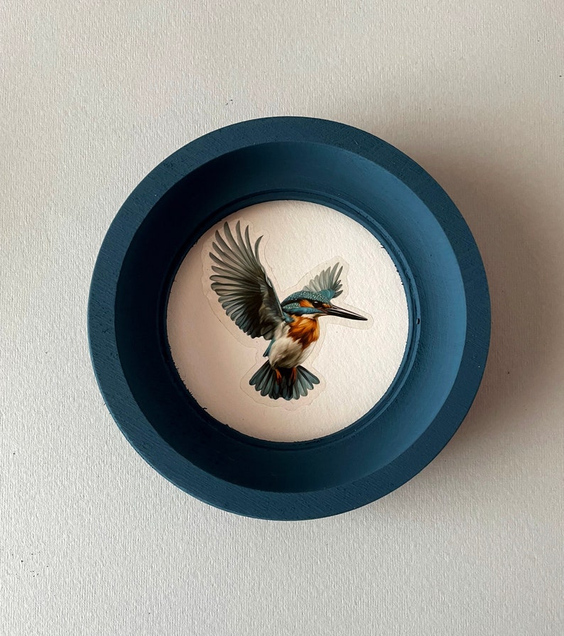 May include: A blue circular frame with a white background and a painting of a blue and brown kingfisher in flight.