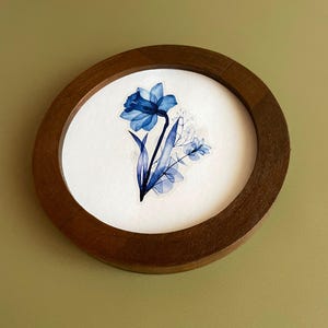 Pode incluir: Uma pintura em aquarela de um narciso azul e outras flores azuis está emoldurada em uma moldura redonda de madeira. A obra de arte está em um fundo branco. A moldura é marrom escuro e tem um acabamento liso. O tema geral é arte floral.