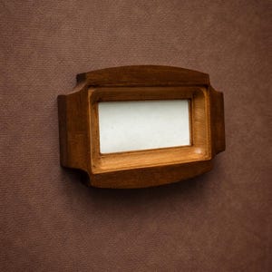 May include: A rectangular wooden nameplate with a light-colored insert. The nameplate has a dark brown finish and is mounted on a textured, brown surface. The design features a slightly curved shape with decorative edges.