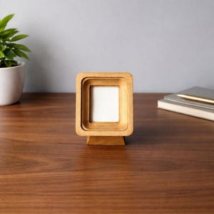 May include: A small, square wooden picture frame with a layered design. The frame is made of light-colored wood and has a white interior. It stands on a matching wooden base. The frame is displayed on a dark wood surface.
