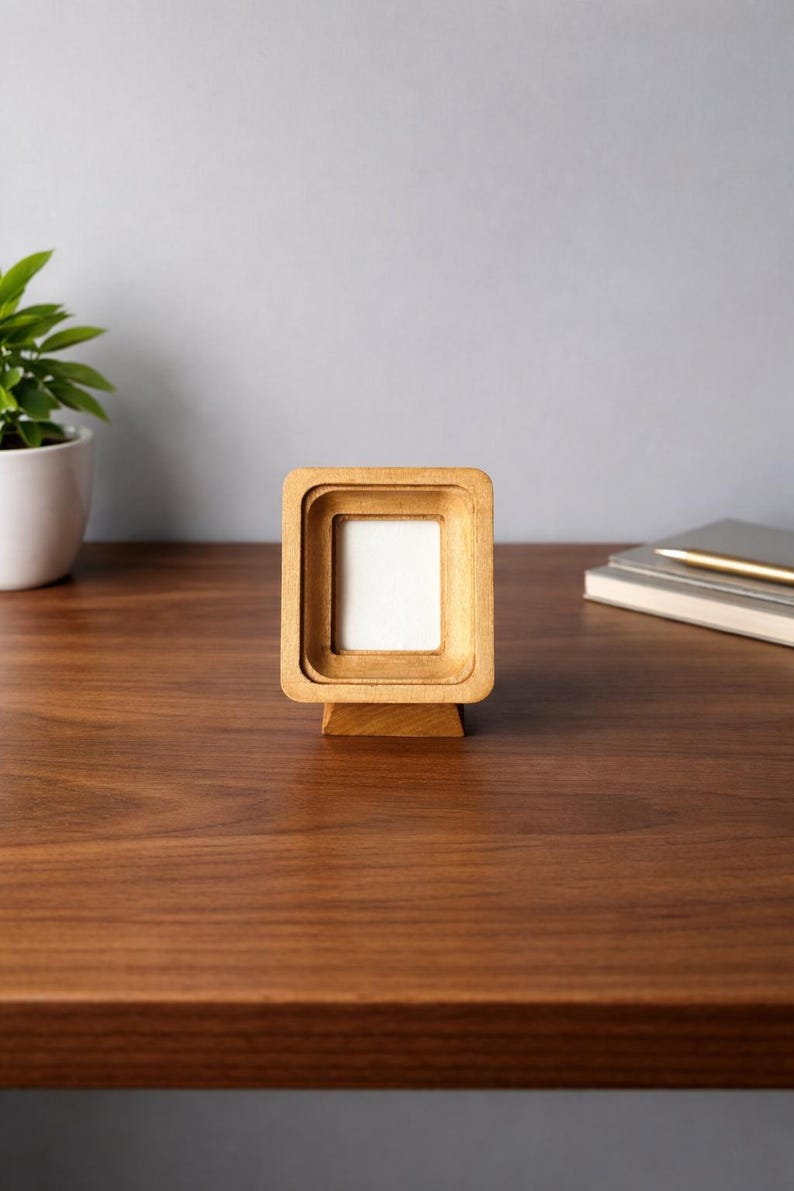 May include: A small, square wooden picture frame with a layered design. The frame is made of light-colored wood and has a white interior. It stands on a matching wooden base. The frame is displayed on a dark wood surface.