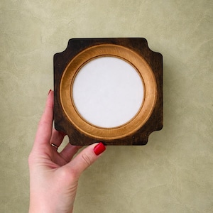 May include: A square wooden frame with a circular opening. The frame has a dark brown exterior and a lighter brown interior ring. The center is white. The frame is held by a hand with red nail polish.