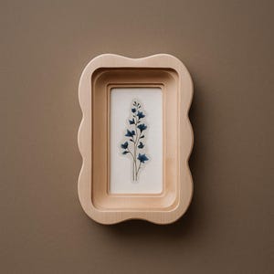 Può includere: Una cornice in legno chiaro con un bordo ondulato, contenente una stampa botanica di fiori blu su sfondo bianco. La cornice ha un design a strati, creando profondità. L'estetica generale è minimalista e naturale.