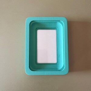 May include: A rectangular picture frame with a teal painted finish. The frame has a recessed inner border and a white interior space for a photo. The frame is set against a neutral background.
