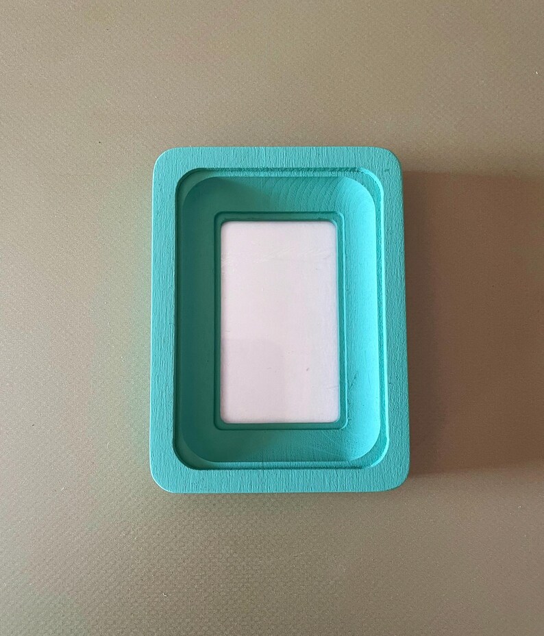 May include: A rectangular picture frame with a teal painted finish. The frame has a recessed inner border and a white interior space for a photo. The frame is set against a neutral background.