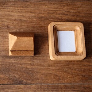 May include: Two wooden items on a wood surface. One is a small, square, light-brown frame with a white insert. The other is a small, light-brown, square block with a pyramid-shaped top. The wood grain is visible.