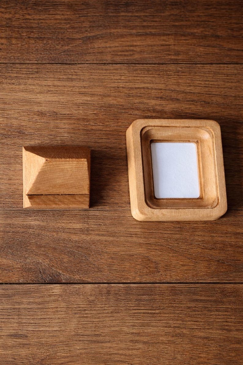 May include: Two wooden items on a wood surface. One is a small, square, light-brown frame with a white insert. The other is a small, light-brown, square block with a pyramid-shaped top. The wood grain is visible.