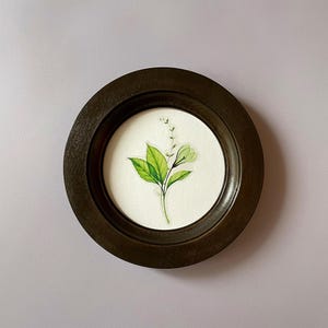 May include: A round, dark brown wooden frame encases a watercolor botanical print. The print features a sprig with vibrant green leaves and delicate white flowers against a white background. The frame has a simple, classic design.