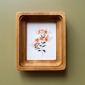 Op de afbeelding: Een klein, houten kunstwerk met een botanische illustratie van oranje lelies met groene bladeren. De lijst is lichtbruin van kleur en heeft een gelaagd ontwerp. Het kunstwerk staat op een witte achtergrond.