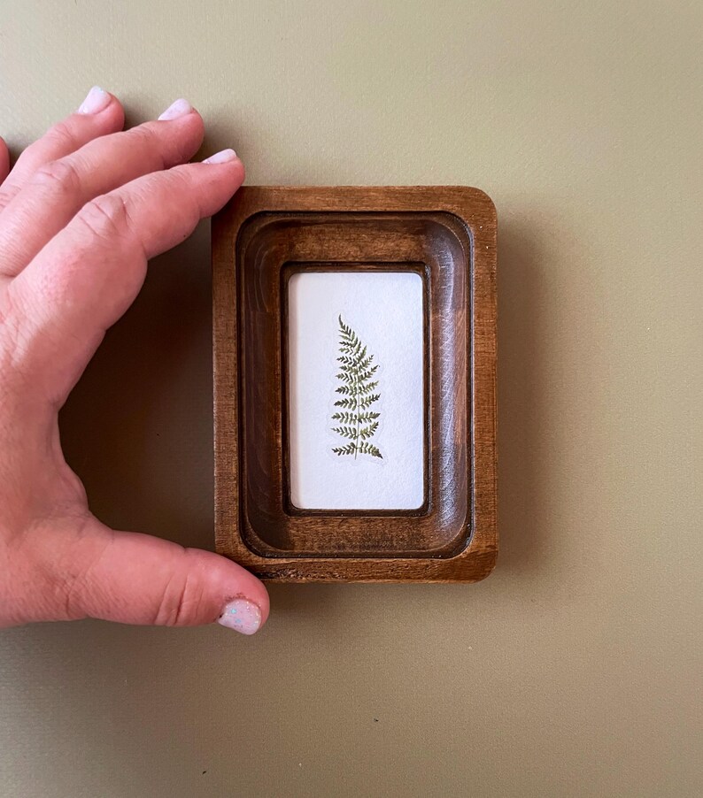 May include: A small, rectangular wooden frame with a recessed design. Inside, a white rectangle displays a delicate fern frond. The frame is held by a hand, showcasing the natural wood grain and the botanical art piece.
