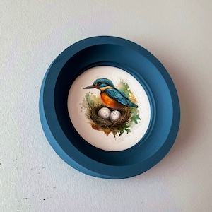 Pode incluir: Um pássaro azul e laranja, o martinho-pescador, sentado em um ninho com três ovos brancos. O pássaro está empoleirado em um galho com folhas verdes. A imagem é emoldurada em uma moldura redonda azul escura.