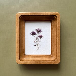 May include: A small, rectangular wooden frame with a light brown finish. Inside the frame is a white background with a cluster of dark purple flowers. The frame is set against a muted green background.
