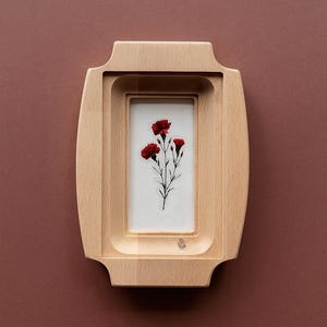 May include: A wooden picture frame with a rectangular opening, showcasing a botanical print of red carnations on a white background. The frame has a light wood finish and a unique, layered design. The background is a solid, muted reddish-brown.