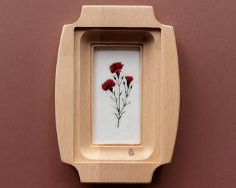 May include: A wooden picture frame with a rectangular opening, showcasing a botanical print of red carnations on a white background. The frame has a light wood finish and a unique, layered design. The background is a solid, muted reddish-brown.