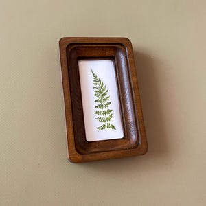May include: A small, rectangular wooden frame with a dark brown finish. Inside the frame is a white background with a green fern pressed against it. The frame is slightly angled, showcasing the natural elements.