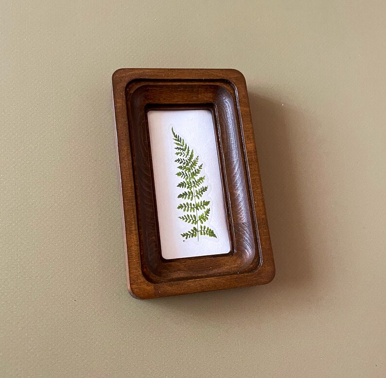 May include: A small, rectangular wooden frame with a dark brown finish. Inside the frame is a white background with a green fern pressed against it. The frame is slightly angled, showcasing the natural elements.