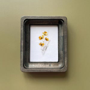 May include: A rectangular wooden frame with a dark, weathered finish containing a white insert. Centered on the white background is a delicate, detailed illustration of a small cluster of yellow flowers with thin green stems and fine foliage.