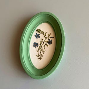 Può includere: Una cornice ovale verde con un'illustrazione botanica di fiori blu e foglie verdi. L'opera d'arte è ambientata su uno sfondo color crema.