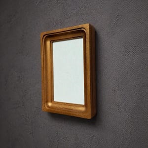May include: A rectangular picture frame with a metallic gold finish and a deep, recessed profile. The frame features a smooth, solid construction and surrounds a blank, white rectangular center area, presented against a dark gray textured wall.