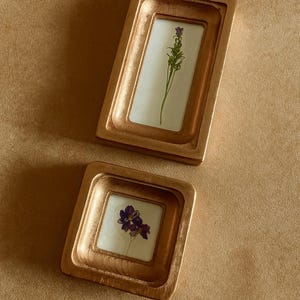 Puede incluir: Dos marcos de fotos rectangulares dorados con flores prensadas en su interior. Un marco es vertical, el otro horizontal. Los marcos están sobre una superficie de color beige.