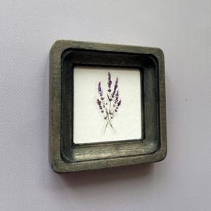 May include: Small, square wooden frame with a distressed gray finish. The frame holds a white background with a sticker of purple lavender sprigs. The frame is approximately 3 inches square.