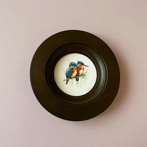 Pode incluir: Uma obra de arte redonda com moldura castanha escura com uma pintura em aquarela de dois guarda-rios empoleirados num galho. As aves têm plumagem azul, laranja e branca. A obra de arte está sobre um fundo branco.