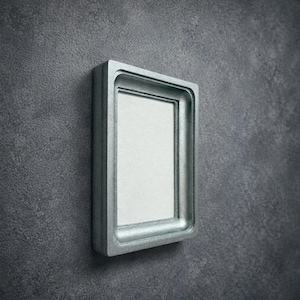 Puede incluir: Un marco de fotos rectangular con un borde grueso de color plata metálica y un panel interior liso de color gris claro. El marco presenta un perfil profundo y escalonado que crea profundidad, sobre un fondo gris oscuro texturizado.