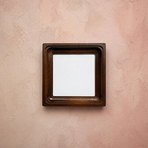 May include: A square wooden frame with a dark brown finish and a smooth, rounded inner edge. The frame surrounds a plain, white, square center panel, creating a clean and simple display piece for mounting or framing various items.