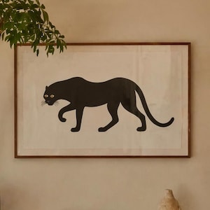 Op de afbeelding: Een ingelijste print van een zwart panter silhouet hangt boven een houten bank. Links staat een potplant. De bank heeft boeken met kleurrijke ruggen en een decoratieve vaas. De kamer heeft een neutraal kleurenpalet.