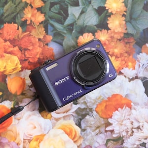 May include: A purple Sony Cybershot digital camera with a black lens, set against a floral backdrop of orange, yellow, and white flowers. The camera displays the Sony and Cybershot logos.