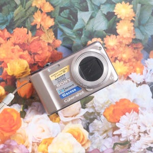May include: A silver Sony Cyber-shot digital camera with a black lens, resting on a floral background of orange, yellow, and white flowers. The camera has a wrist strap attached and several stickers.