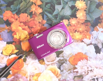 May include: A magenta Nikon Coolpix digital camera featuring a silver lens barrel, a silver shutter button, and a black wrist strap attached to the side. The camera body displays the Nikon brand name and the Coolpix model name in white text.