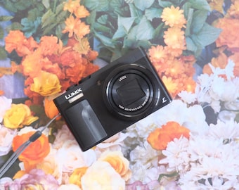 May include: A black Panasonic Lumix digital camera featuring a Leica lens with a retractable cover. The camera body includes a textured grip on the front, a silver wrist strap attached to the side, and the Lumix brand name printed in white.