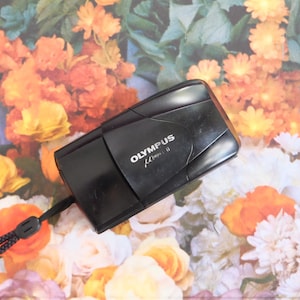May include: A black Olympus Mju II 35mm film camera with a sliding lens cover and a black wrist strap. The camera body features the white text Olympus Mju II printed on the front surface. The device is compact with a smooth, rounded rectangular shape.