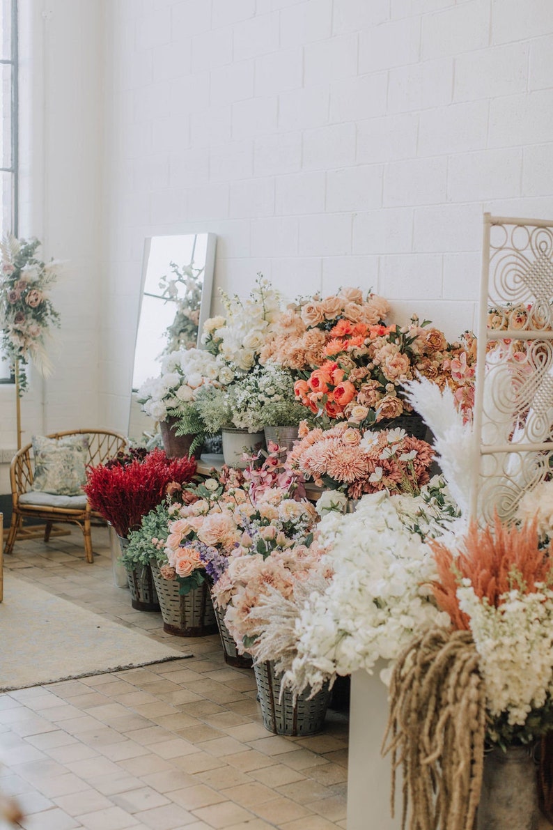 May include: A variety of dried flowers and greenery arranged in baskets and containers. The flowers are in shades of pink, white, and red. The flowers are arranged in a row against a white brick wall.