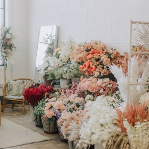 May include: A variety of dried flowers and greenery arranged in baskets and containers. The flowers are in shades of pink, white, and red. The flowers are arranged in a row against a white brick wall.