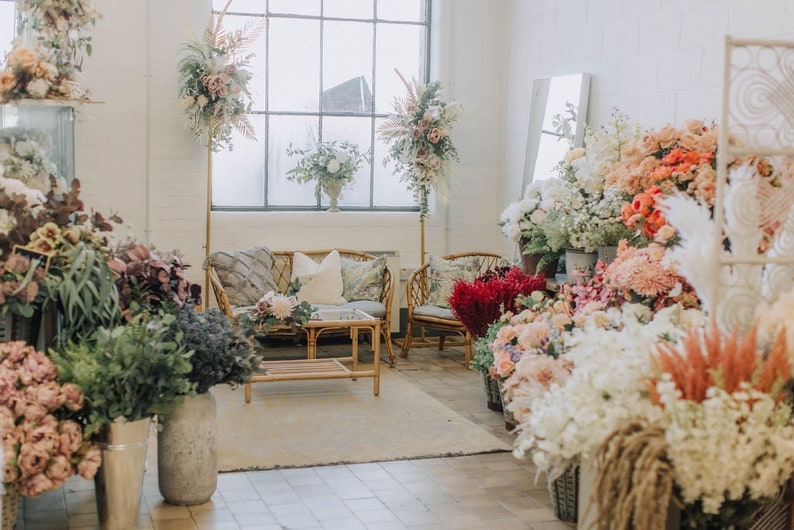 May include: A floral shop interior with a white brick wall, a large window, and a wicker sofa with pillows. There are many bouquets of flowers in various colors and styles, including white, pink, red, and orange.