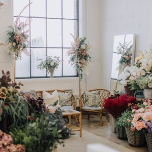 May include: A floral shop interior with a variety of flowers in different colors and arrangements. There are two wicker chairs and a small table in the center of the room. The shop has a large window and a white brick wall.