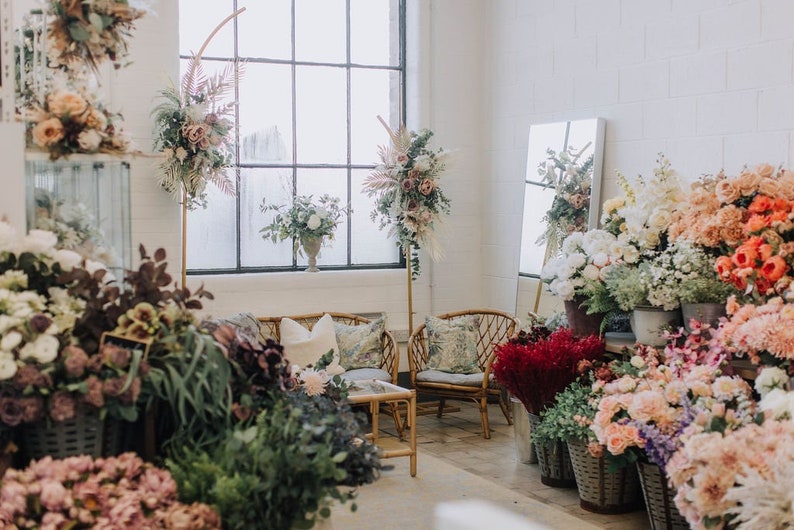 May include: A floral shop interior with a variety of flowers in different colors and arrangements. There are two wicker chairs and a small table in the center of the room. The shop has a large window and a white brick wall.