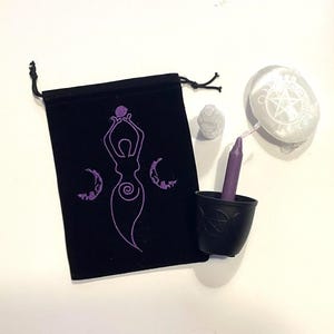 May include: A black velvet pouch with a purple embroidered design of a woman holding a sphere with three crescent moons. The pouch is next to a purple candle in a black holder with a pentagram design.