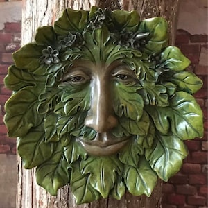 May include: A green man wall sculpture with a serene face surrounded by detailed leaves and small flowers. The sculpture has a bronze-colored face and is mounted on a tree trunk, with a brick wall in the background.
