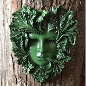 May include: A green Green Man wall hanging. The face is surrounded by a leafy design. The sculpture is a shade of green and is mounted on a wooden surface. The Green Man is a symbol of rebirth and growth.