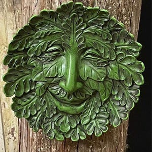 May include: A green, leaf-covered face sculpture with a smiling expression. The face is surrounded by detailed leaves, creating a natural, woodland theme. The sculpture is mounted on a textured, wooden surface.